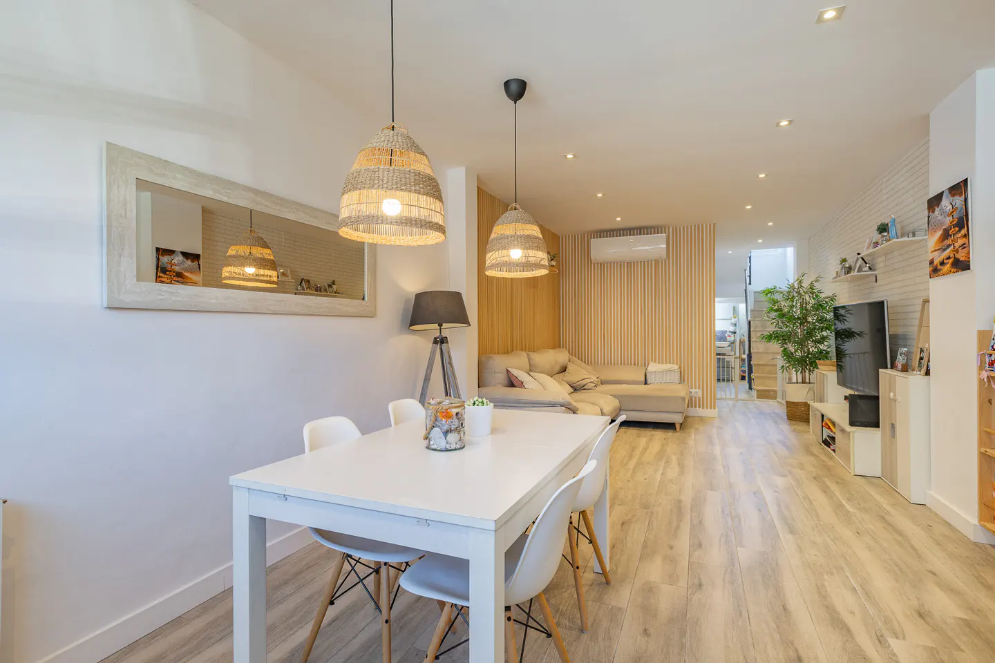 Bright, modern living space with white dining table, chairs, and woven pendant lights. Beige sectional sofa and TV in the background.