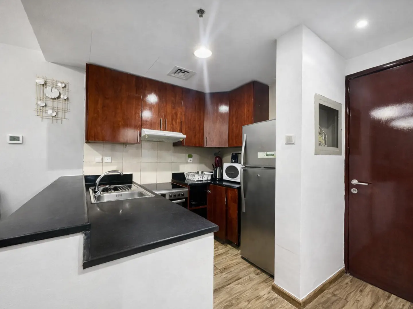 A compact kitchen with dark wood cabinets, black countertops, and stainless steel appliances. A dark brown door is on the right.