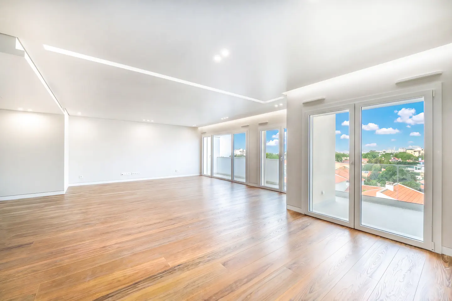Bright, empty room with hardwood floors, white walls, and large windows showing a city view with blue sky.