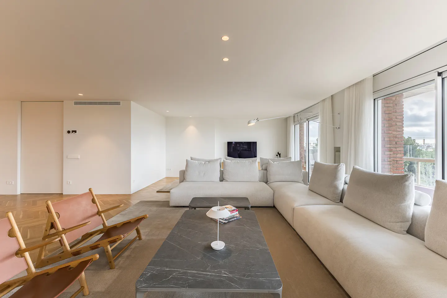 A bright, modern living room with a large white sectional sofa, marble coffee table, and two pink leather chairs. Large windows offer a city view.