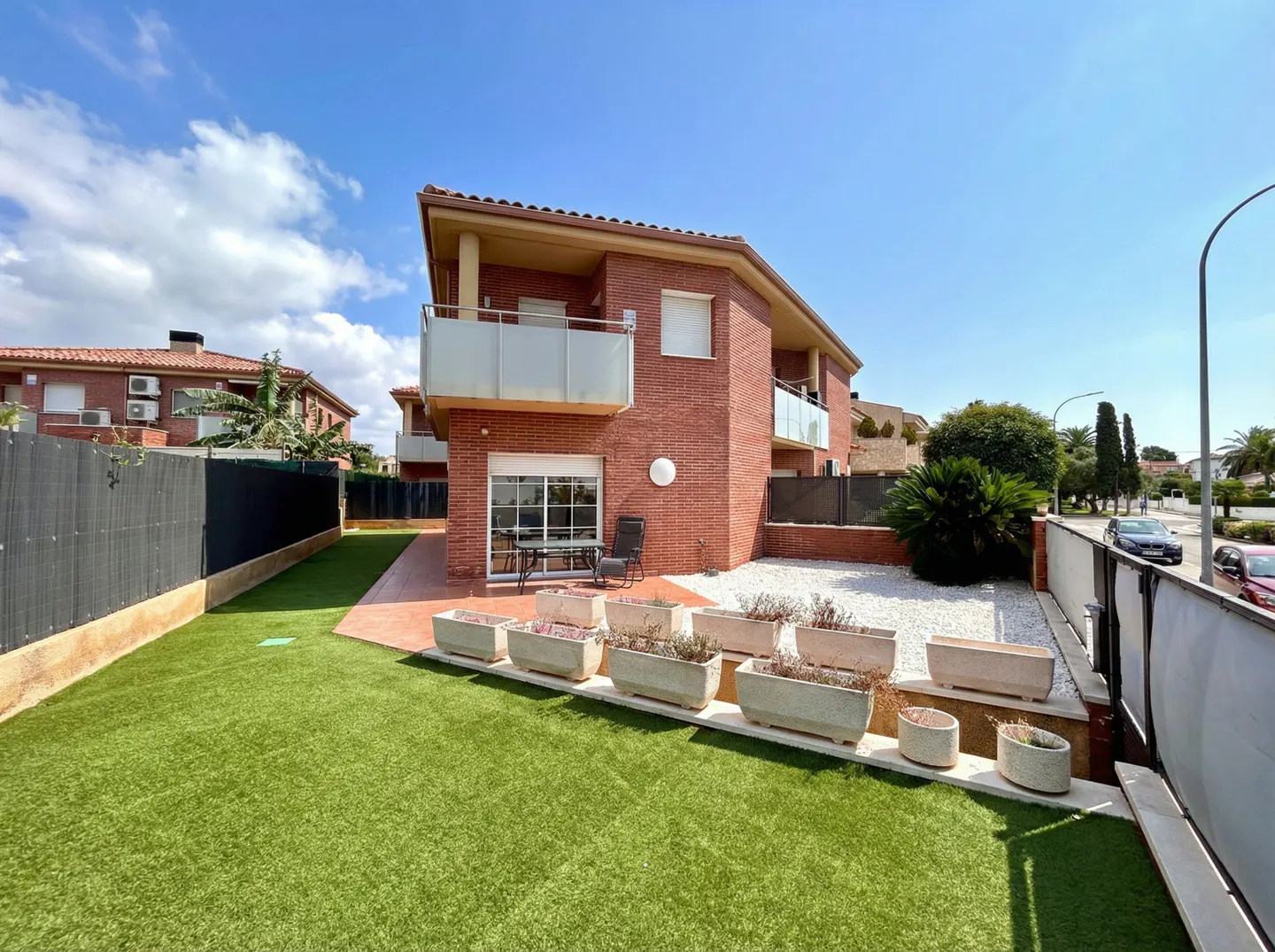 Two-story red brick house with a balcony, green lawn, and potted plants under a blue sky.