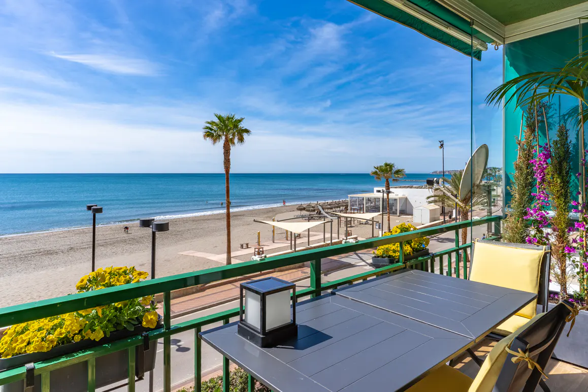 Balcony view with a table and chairs overlooking a sandy beach, palm trees, and the blue ocean under a clear sky.