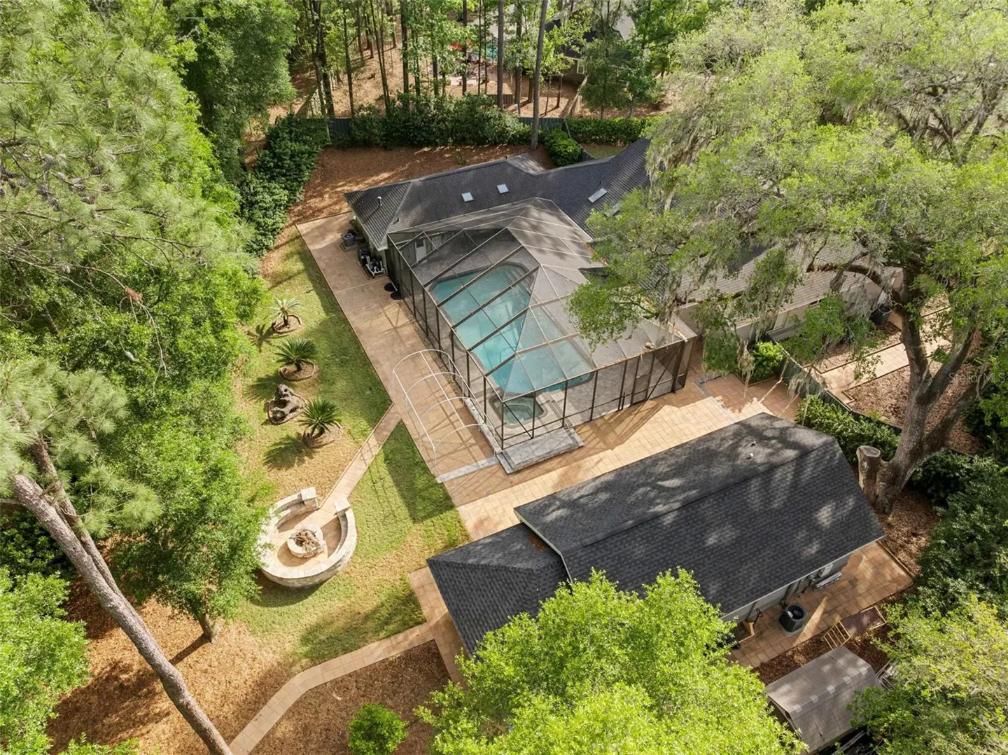 Aerial view of a home with a screened-in pool, fire pit, and lush green trees. The house has a dark roof and light-colored siding.