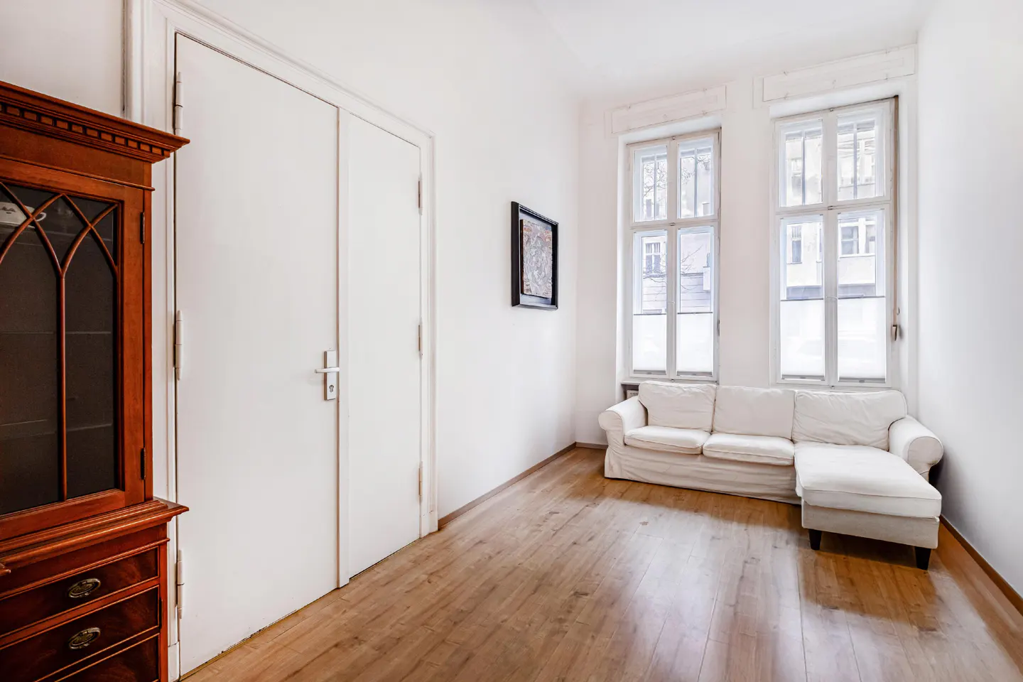 Bright room with wood floors, white walls, and two windows. A white sofa sits beneath the windows. A wooden cabinet is on the left, and a framed picture hangs on the wall.