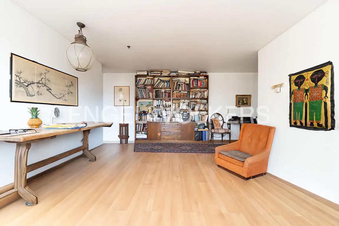 Bright study with light wood floors, a long wooden desk, a large floor-to-ceiling bookshelf, and an orange armchair.