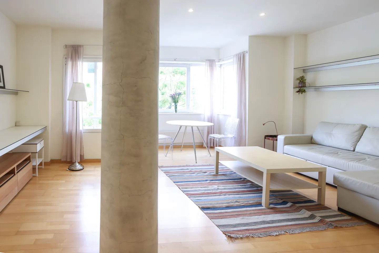 Bright living room with a beige column, light wood floors, white sofa, and a striped rug. A table and chairs are near the window.
