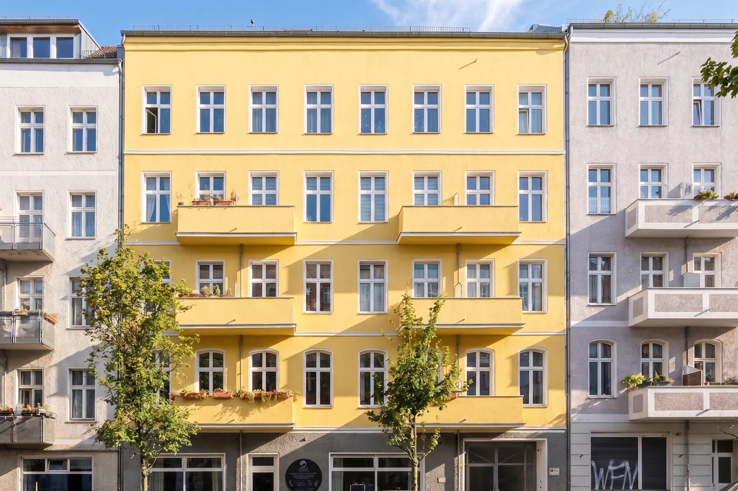 A bright yellow four-story apartment building with balconies, flanked by white and grey buildings on a sunny day.