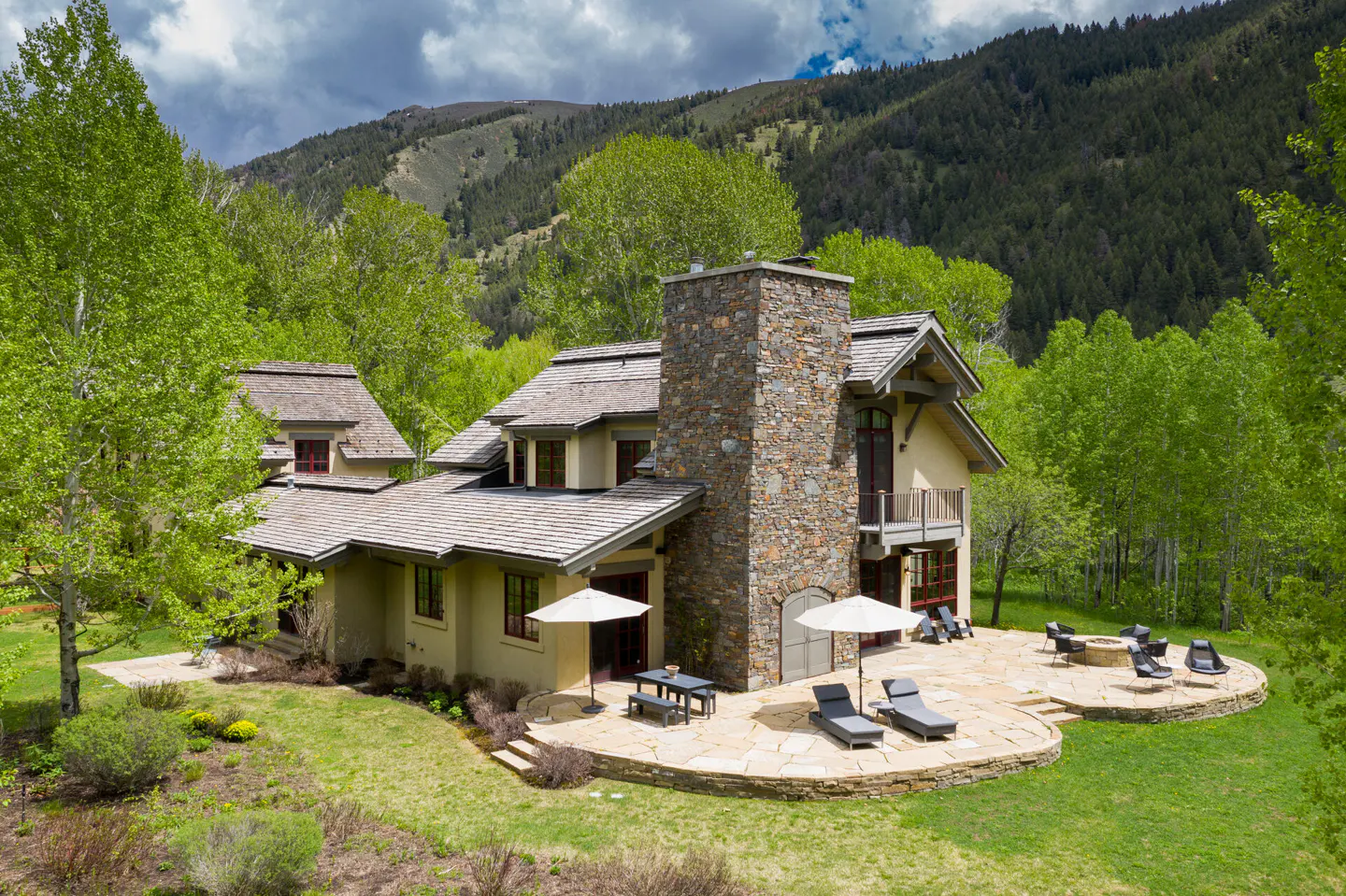 Exterior view of a tan house with a stone chimney, patio, and mountain backdrop.