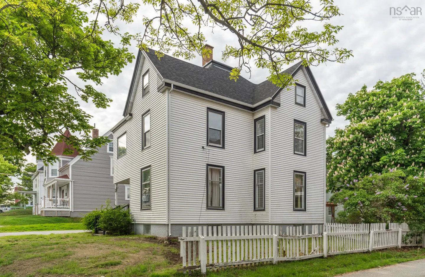 Two-story white house with black trim and a dark roof, surrounded by a white picket fence and lush green trees.
