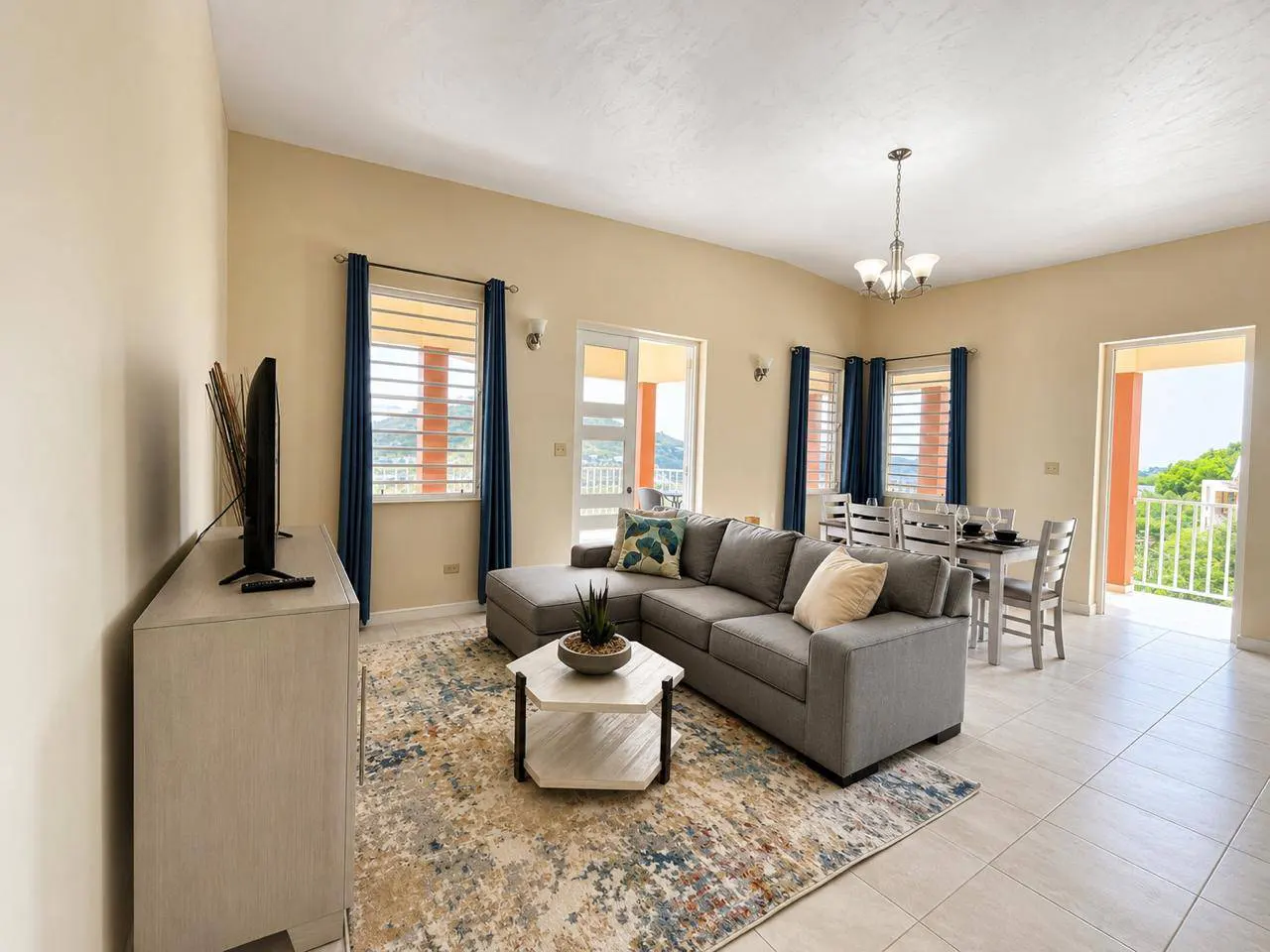 Living room with gray sectional sofa, patterned rug, and dining table. Balcony with a view is visible. Blue curtains frame the windows.