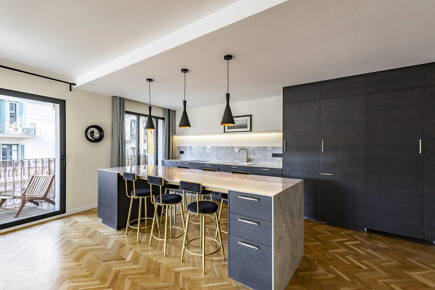 A modern kitchen with a large island, black cabinets, gold stools, and herringbone wood floors. Balcony with a chair is visible.