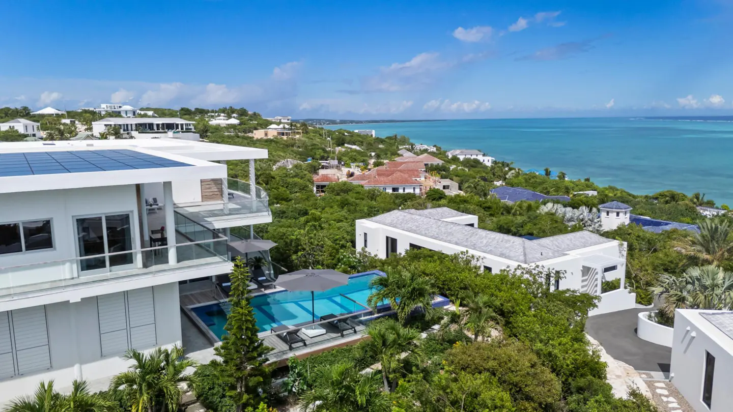 Aerial view of a modern white villa with a pool, solar panels, and ocean views under a blue sky. Lush green trees surround the property.