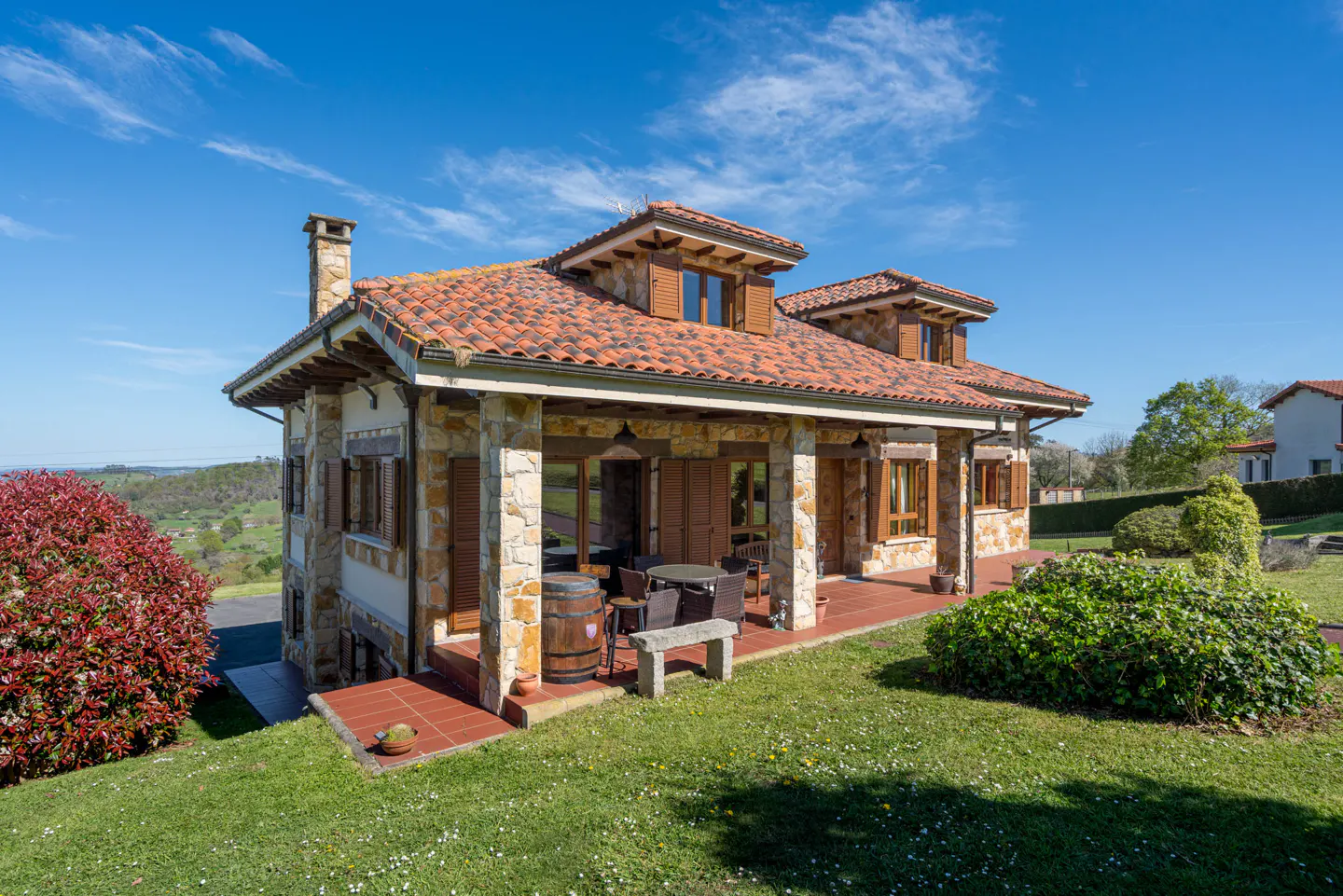 Exterior view of a stone house with a red tile roof, a covered patio with furniture, and a green lawn under a blue sky.