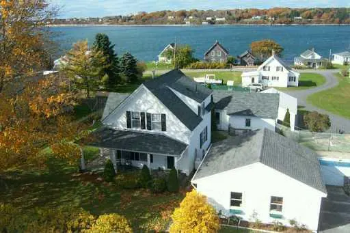 Aerial view of a white house with a dark roof, surrounded by green grass and trees, with a body of water in the background.