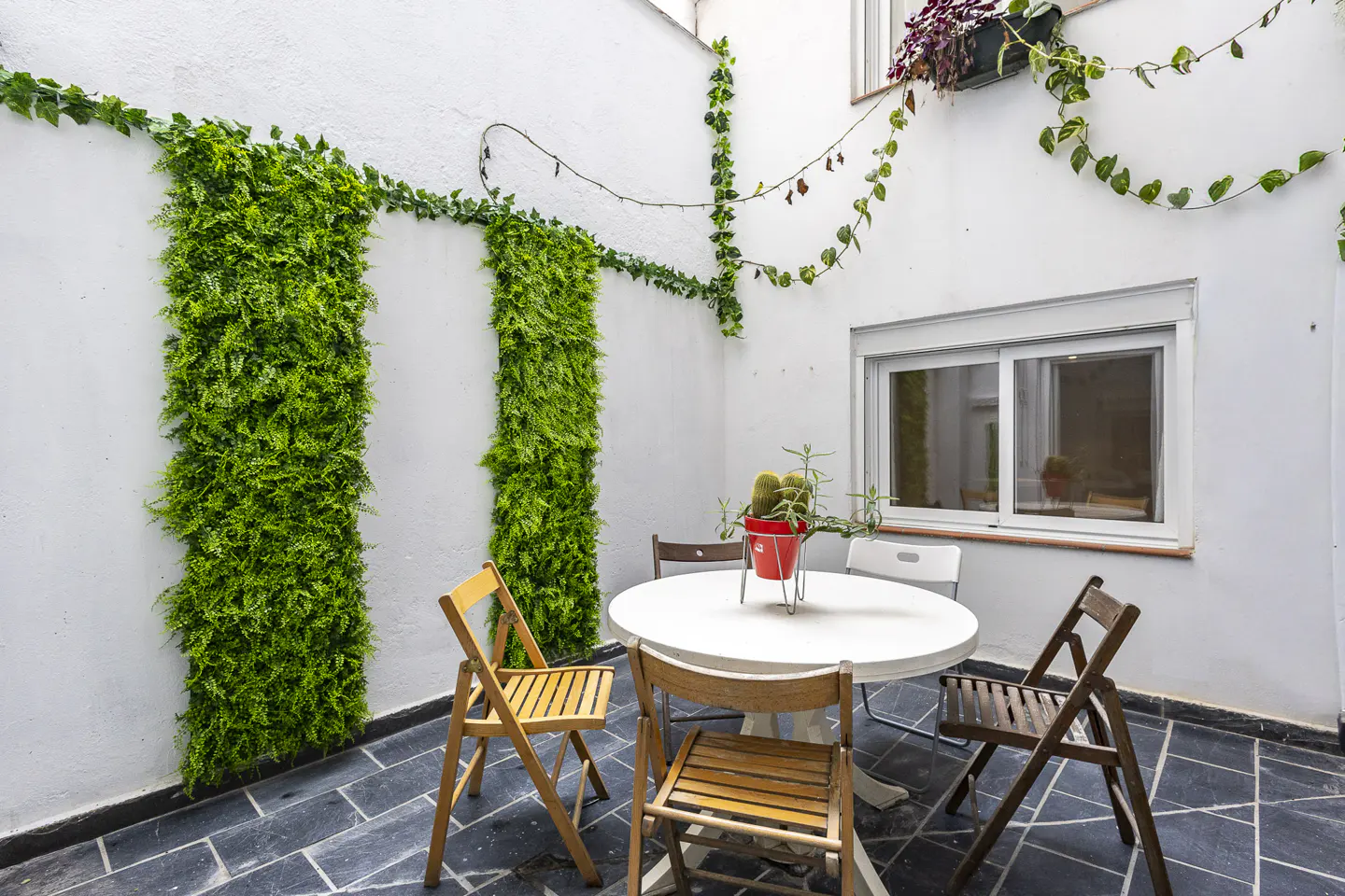 Small patio with a white round table, wooden chairs, and vertical green plant walls against a white exterior wall.