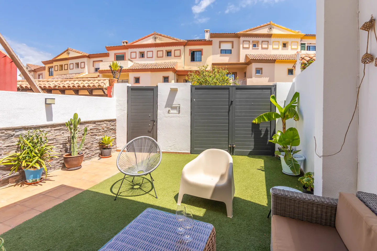 A sunny patio with artificial grass, two chairs, a table, and a sofa. In the background are townhouses with orange roofs and white walls.