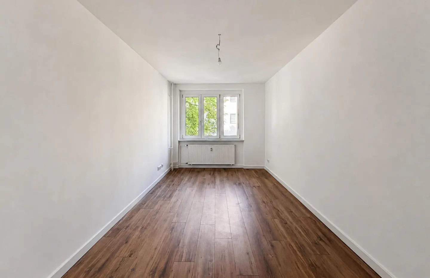 Empty room with white walls, dark wood-look flooring, a central window with a view of trees, and a white radiator below.