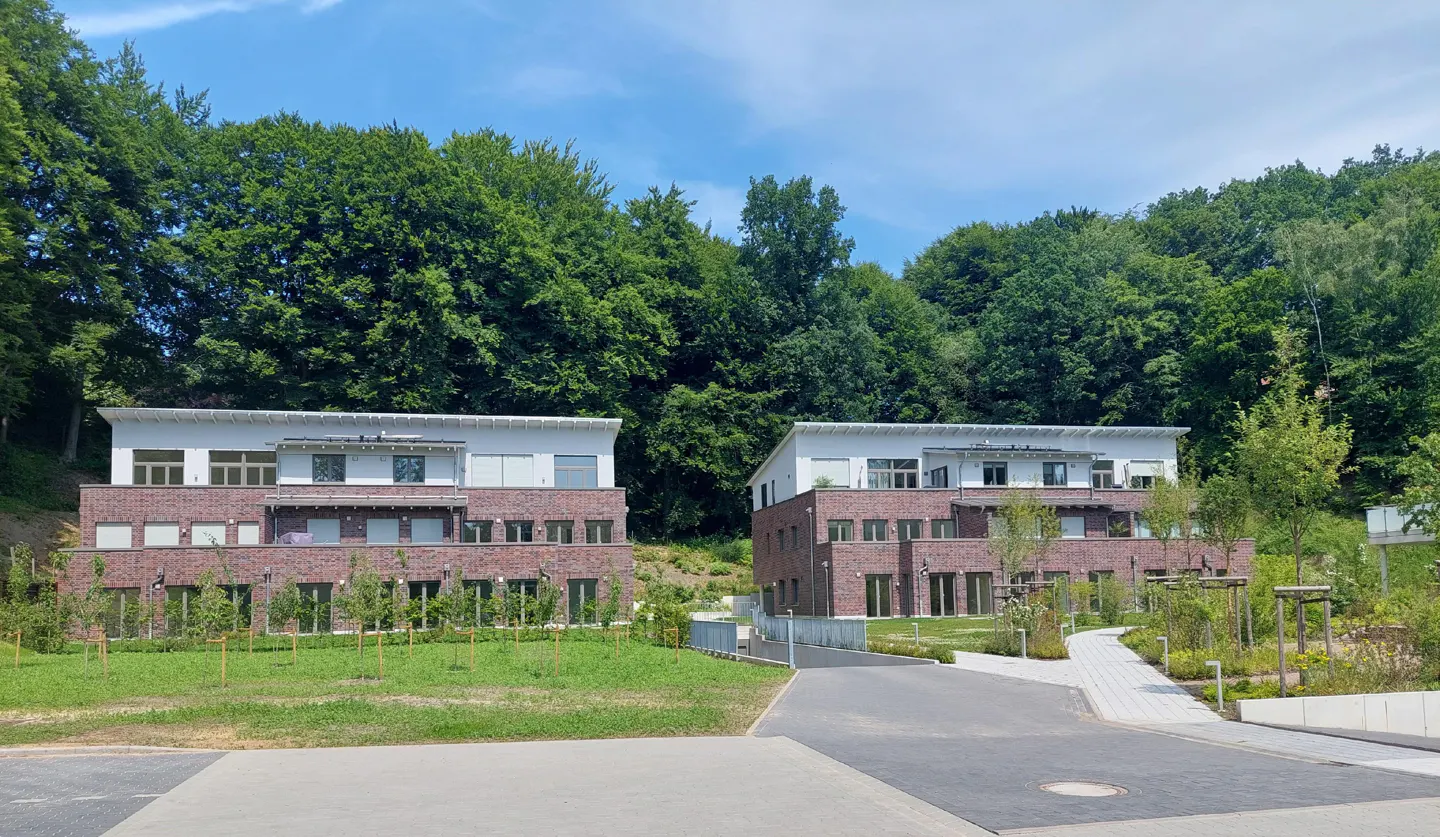 Two modern brick apartments with white roofs sit at the edge of a lush green forest under a bright blue sky.