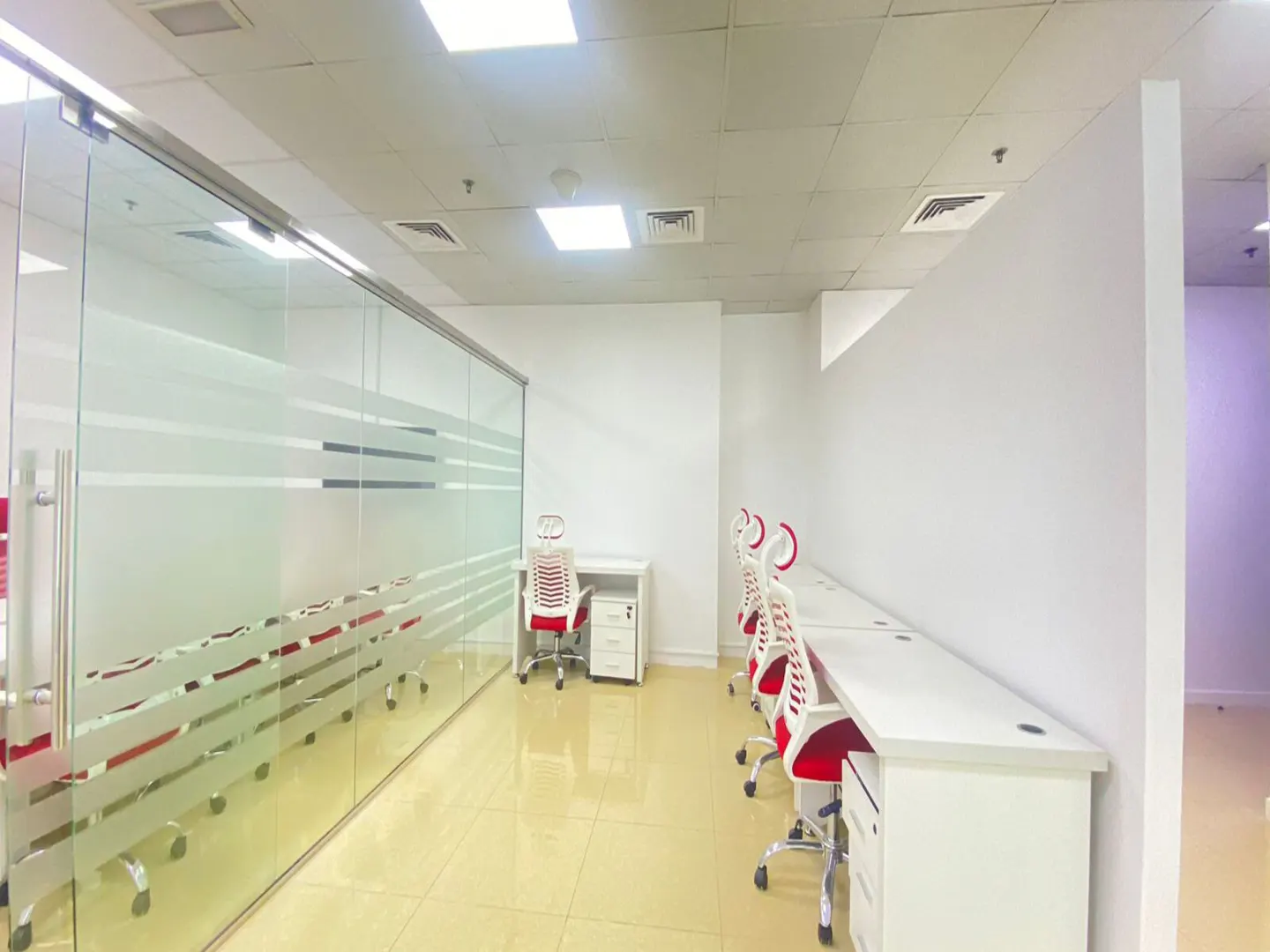 Bright office space with white walls, tan tile floor, and white desks. Red and white chairs line one wall. A glass-walled conference room is on the left.