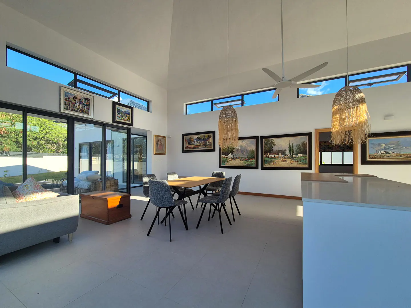 Bright, modern living room with white walls, large windows, and a dining table with six chairs. Paintings adorn the walls. A ceiling fan and pendant lights add to the ambiance.