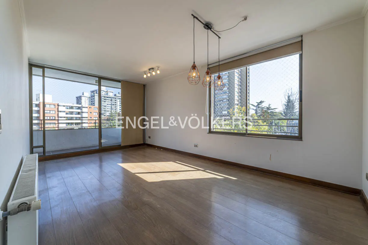 Bright, empty living room with hardwood floors, white walls, and large windows showing city views. Modern pendant lights hang from the ceiling.