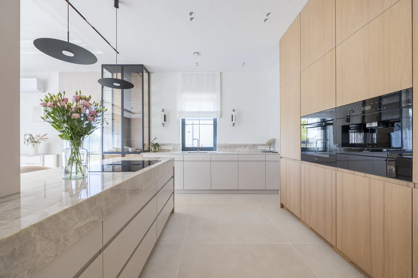 A modern kitchen with light wood cabinets, a marble island with pink flowers, and black pendant lights.