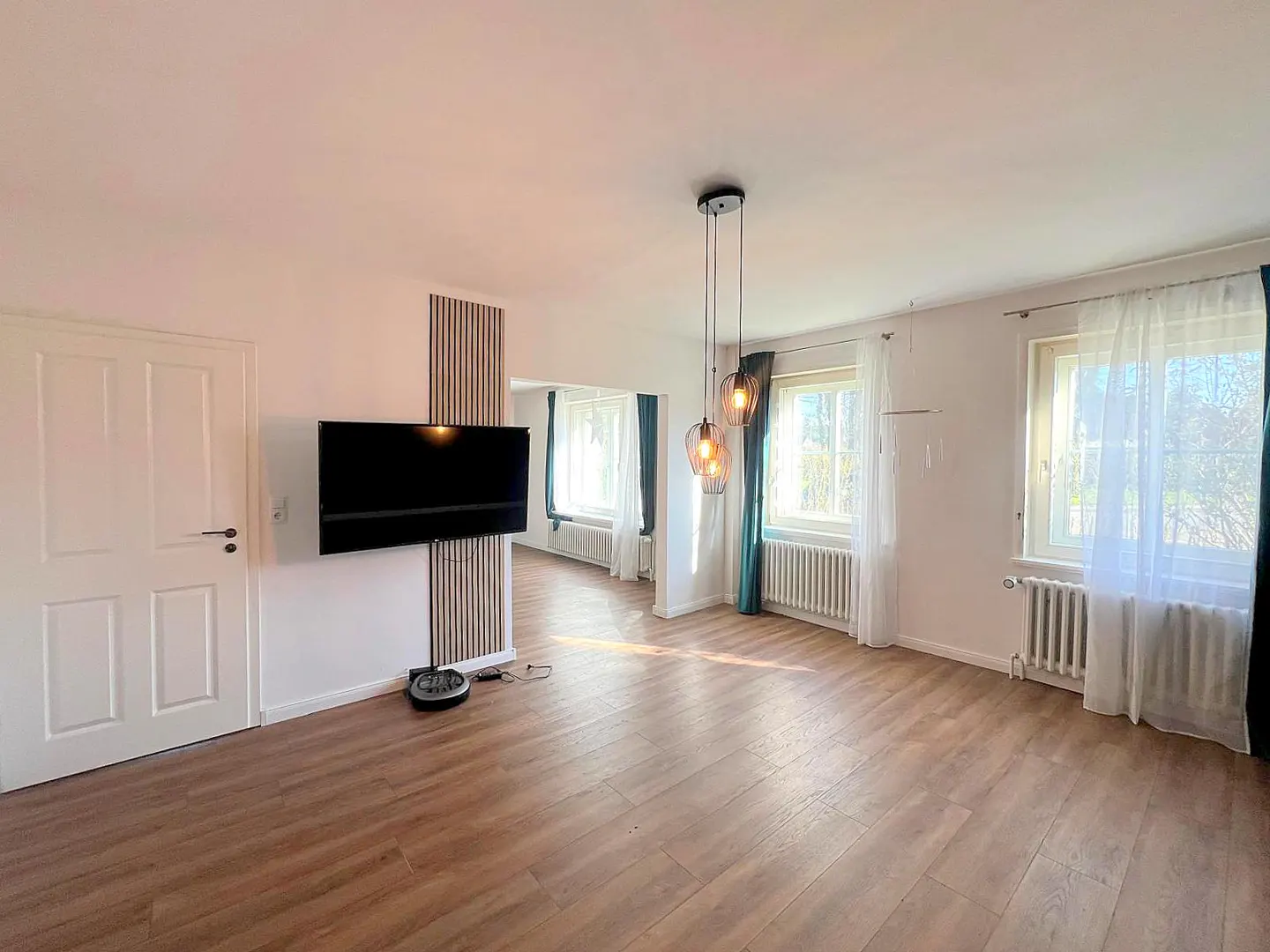 Bright living room with wood floors, white walls, and three windows with sheer curtains. A TV is mounted on a wood-paneled wall.