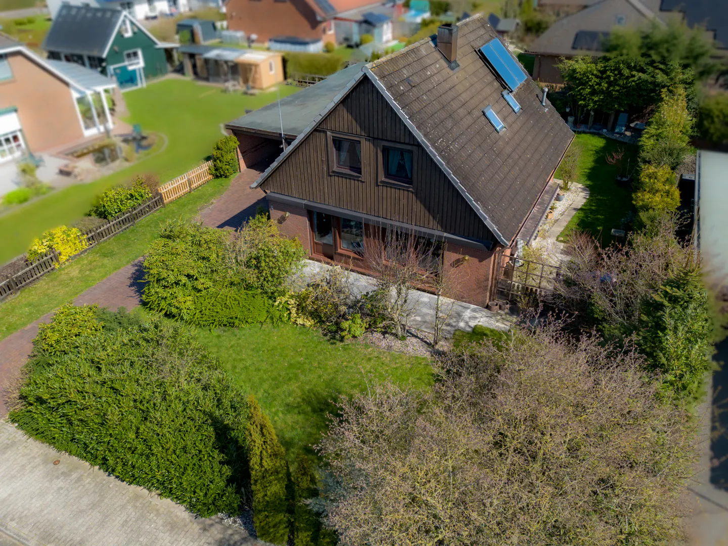 Aerial view of a brown A-frame house with a brick base, a brown tiled roof, solar panels, and a green lawn.
