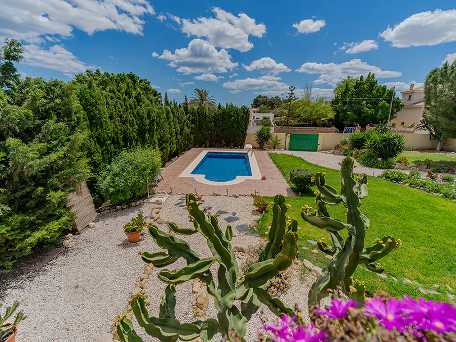 Aerial view of a backyard with a blue swimming pool, green lawn, cacti, and white gravel under a blue sky with white clouds.