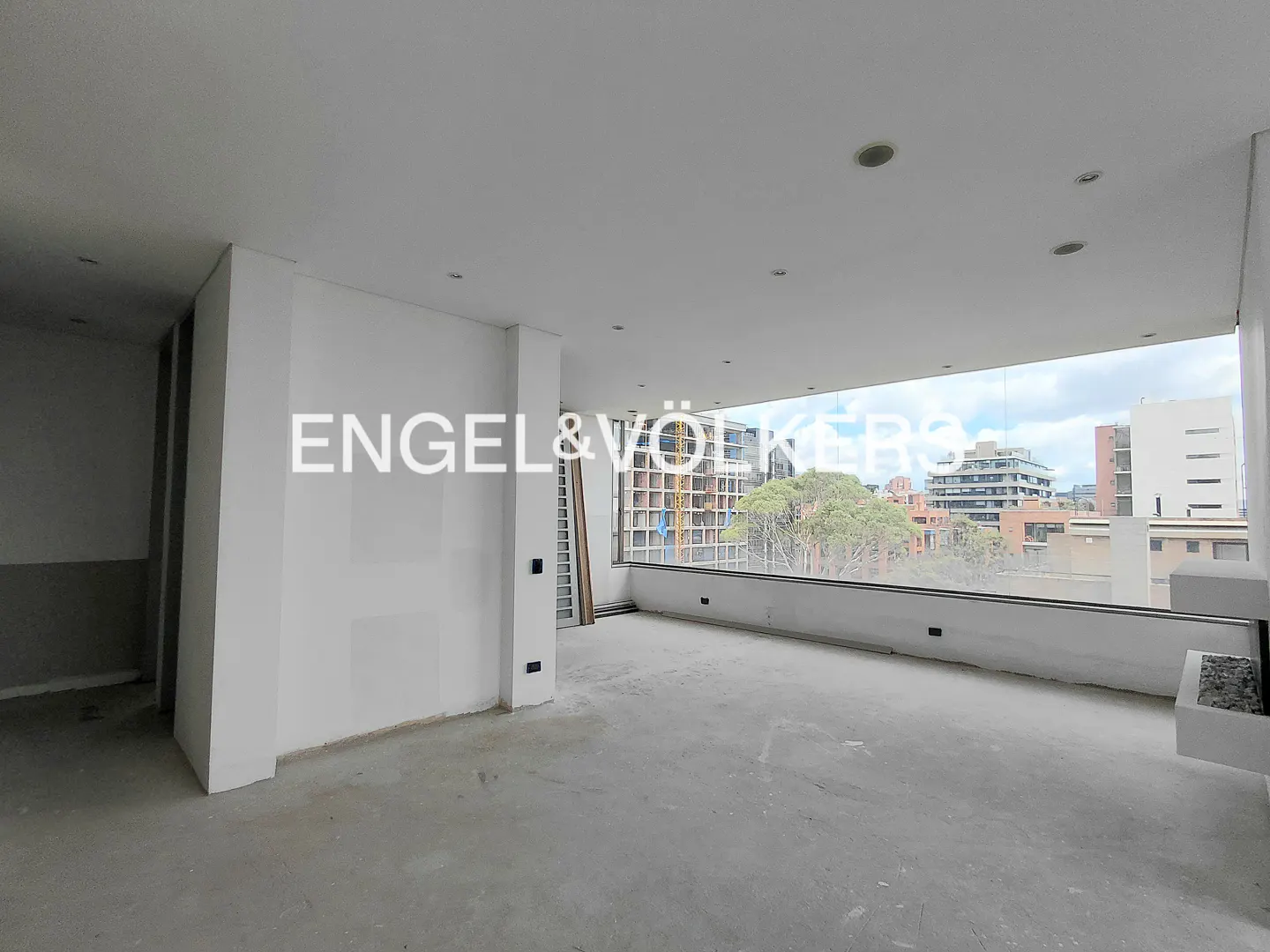 Unfinished room with white walls and concrete floor. Large window with city view. Engel & Völkers logo.