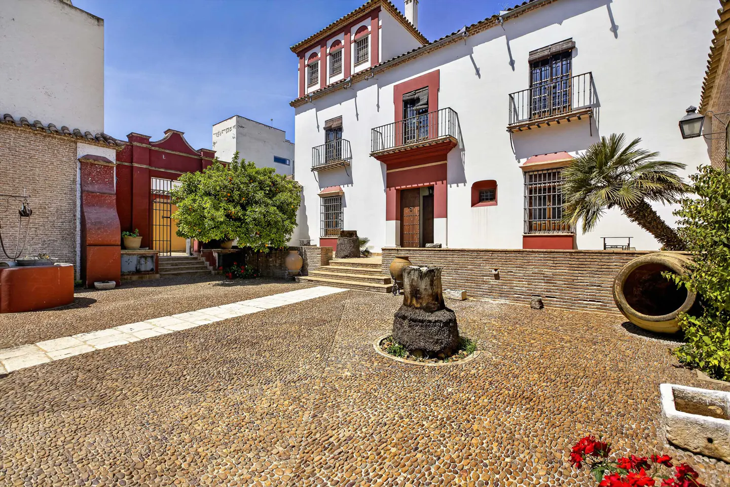 Courtyard view of a two-story white building with red trim and black balconies. The ground is cobblestone with a stone feature in the center.