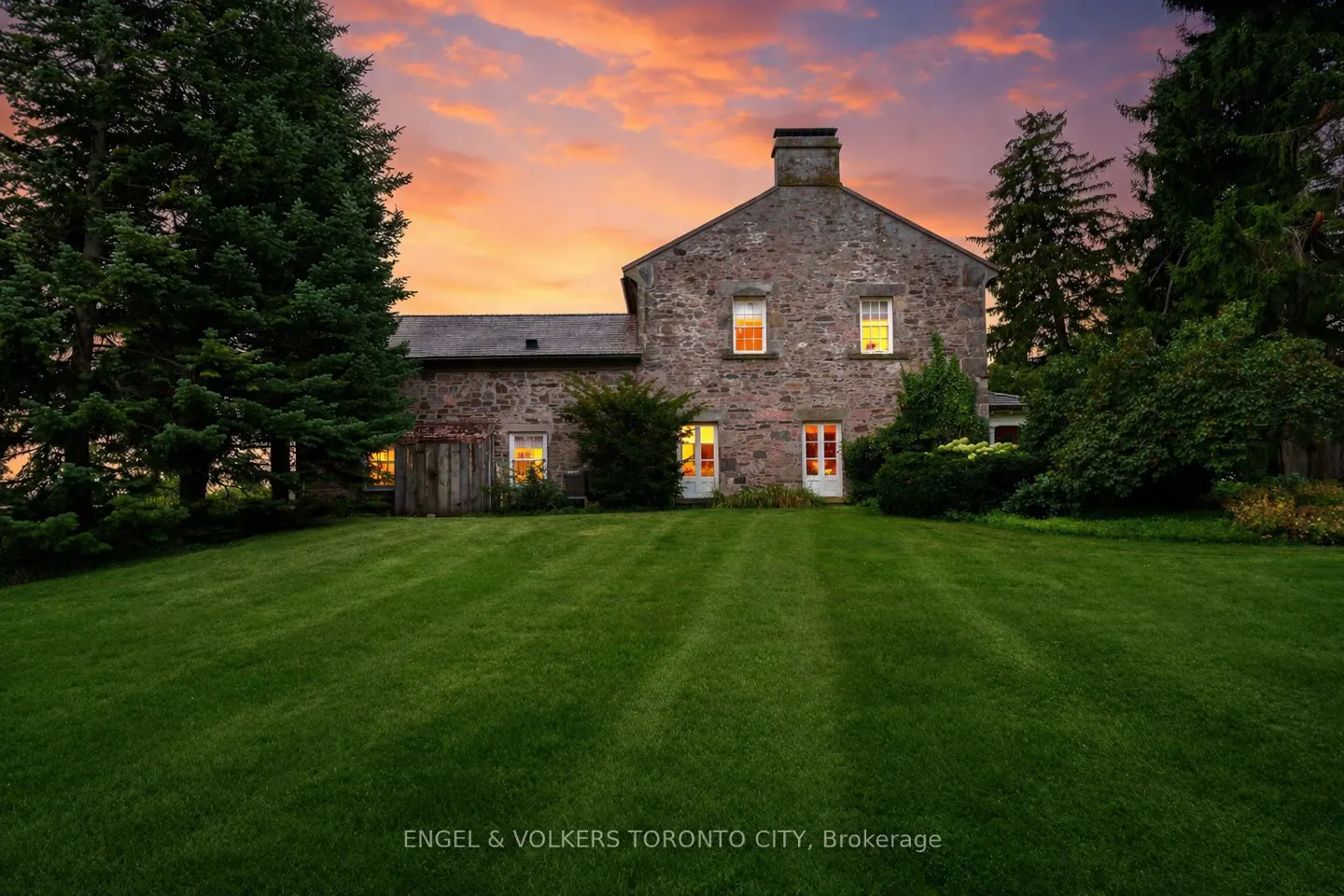 Exterior of a stone house with a green lawn at sunset. Lights are on inside.