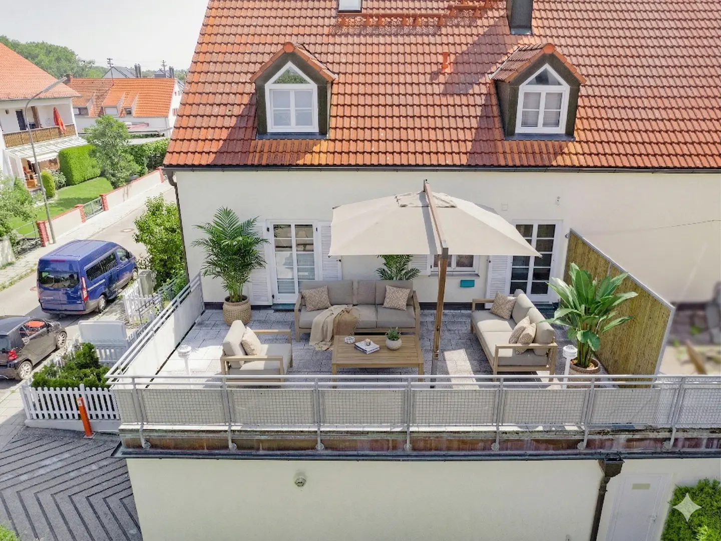 Rooftop patio with beige furniture, umbrella, and plants. White building with red tile roof and dormer windows. Railing in foreground.