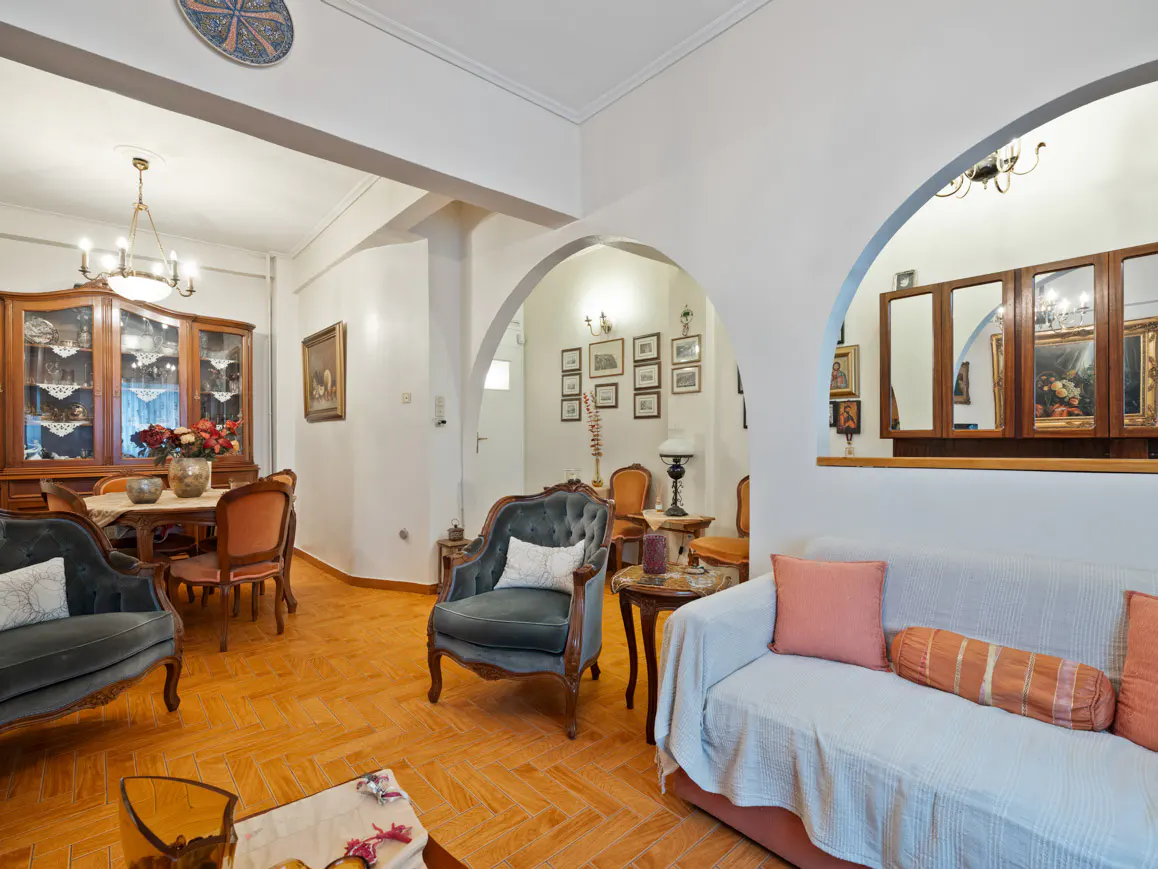 A living room with a sofa, chairs, and a dining table, featuring white walls and arched doorways.