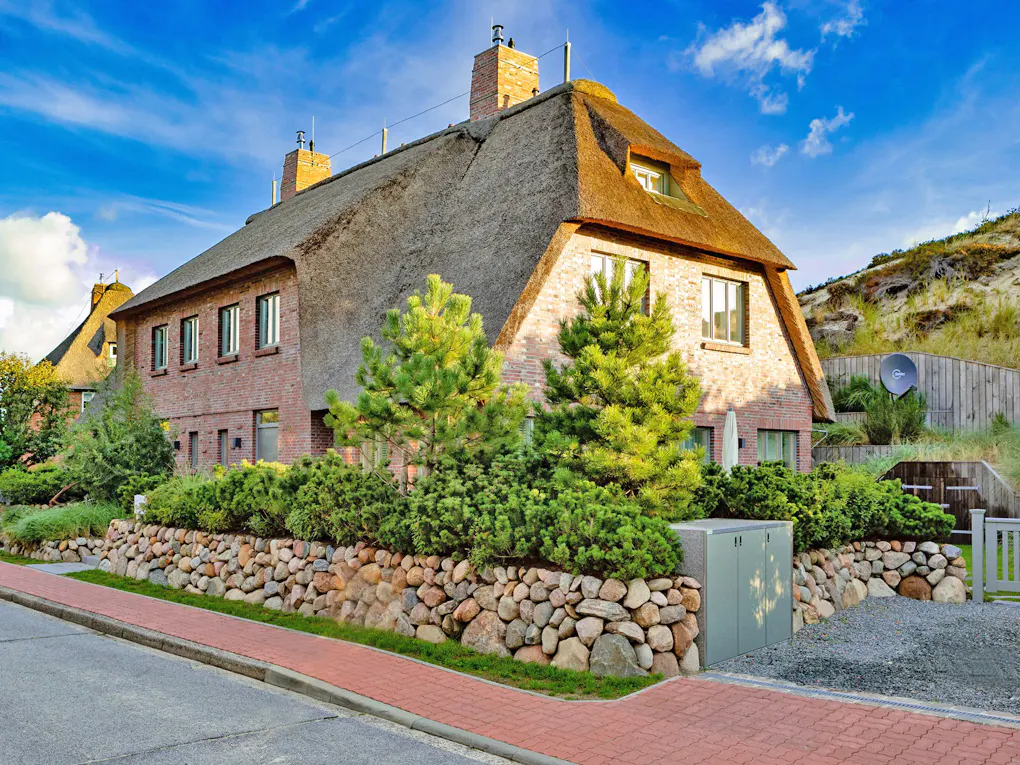 A brick house with a thatched roof sits behind a stone wall and green bushes.