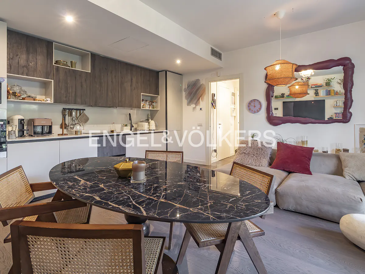 Open-concept living space with a black marble table, rattan chairs, and a gray sofa with a red pillow. Modern kitchen in the background.