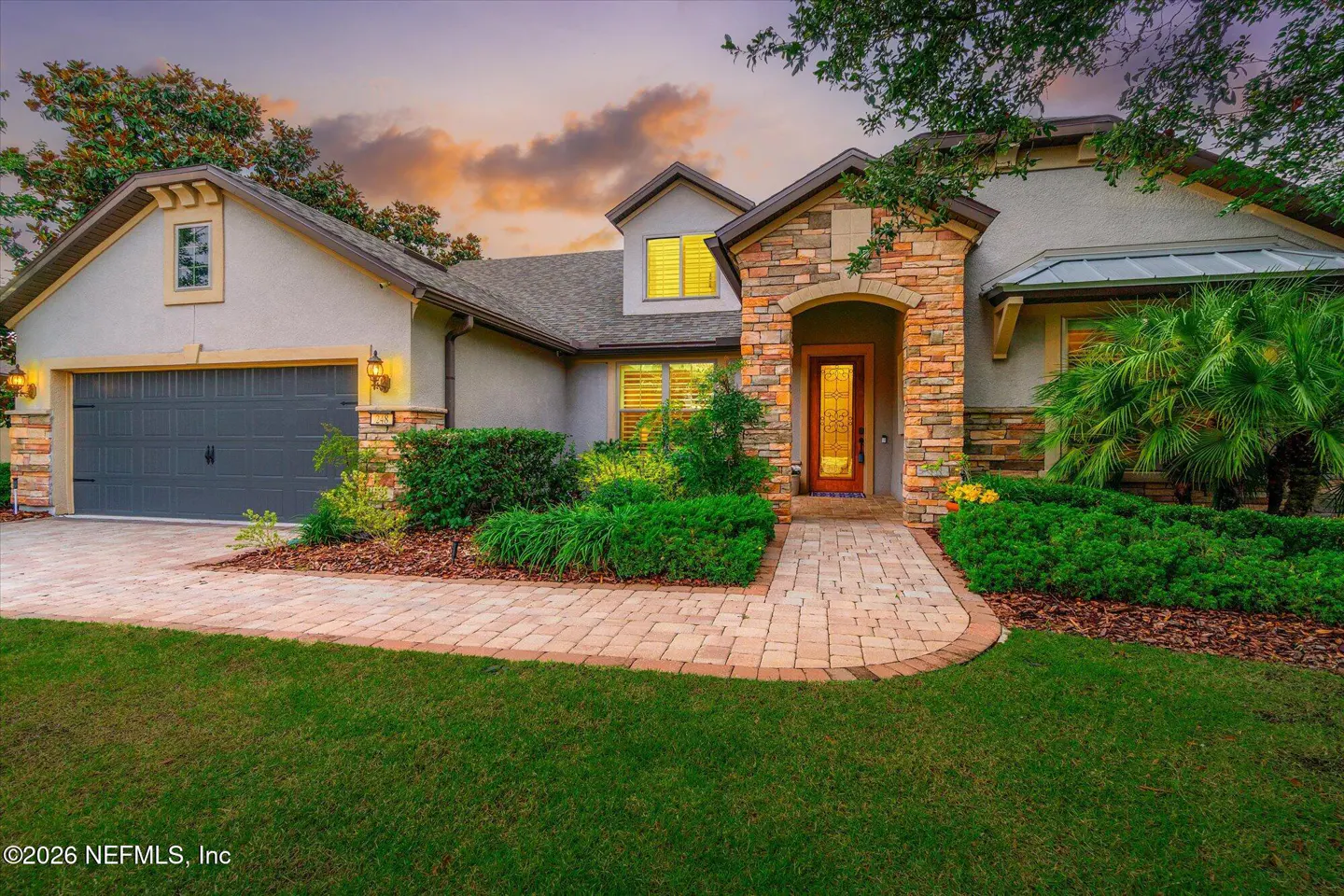 Single-story home with stone accents, a gray garage, and a paver walkway at sunset with lush landscaping.
