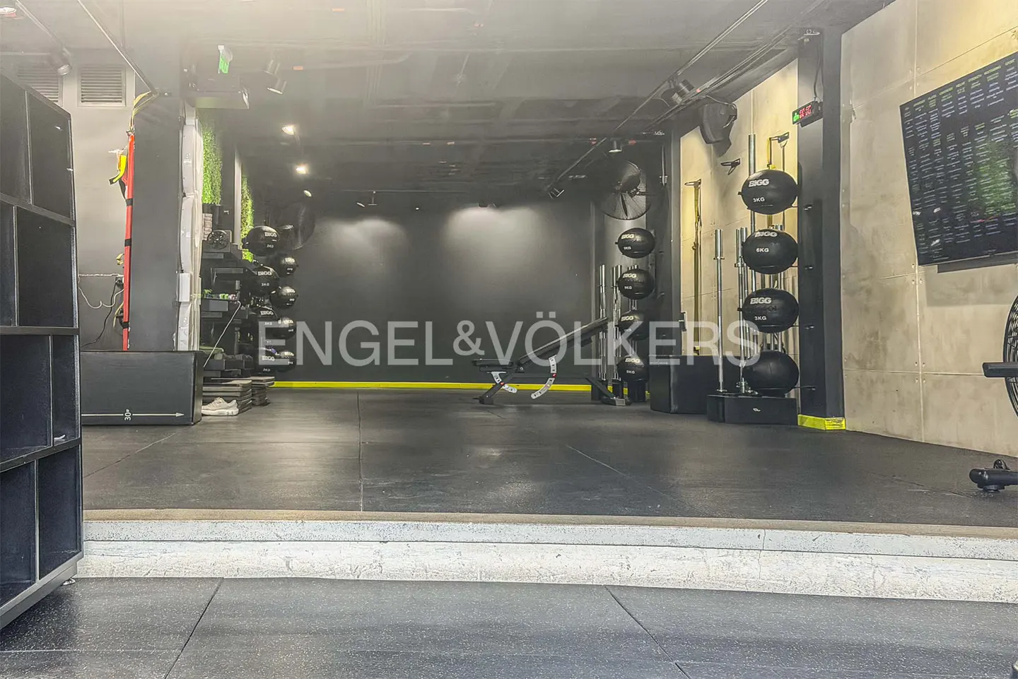 A modern gym with black rubber flooring, weight racks, and an Engel & Völkers logo on the back wall.
