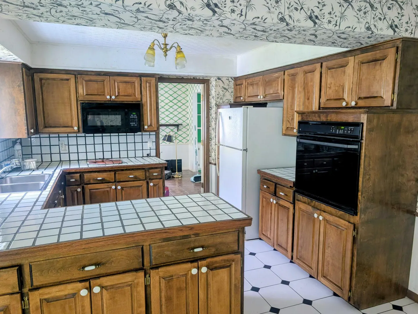 A kitchen with wood cabinets, white appliances, and black and white tile floors. The ceiling has bird-themed wallpaper.