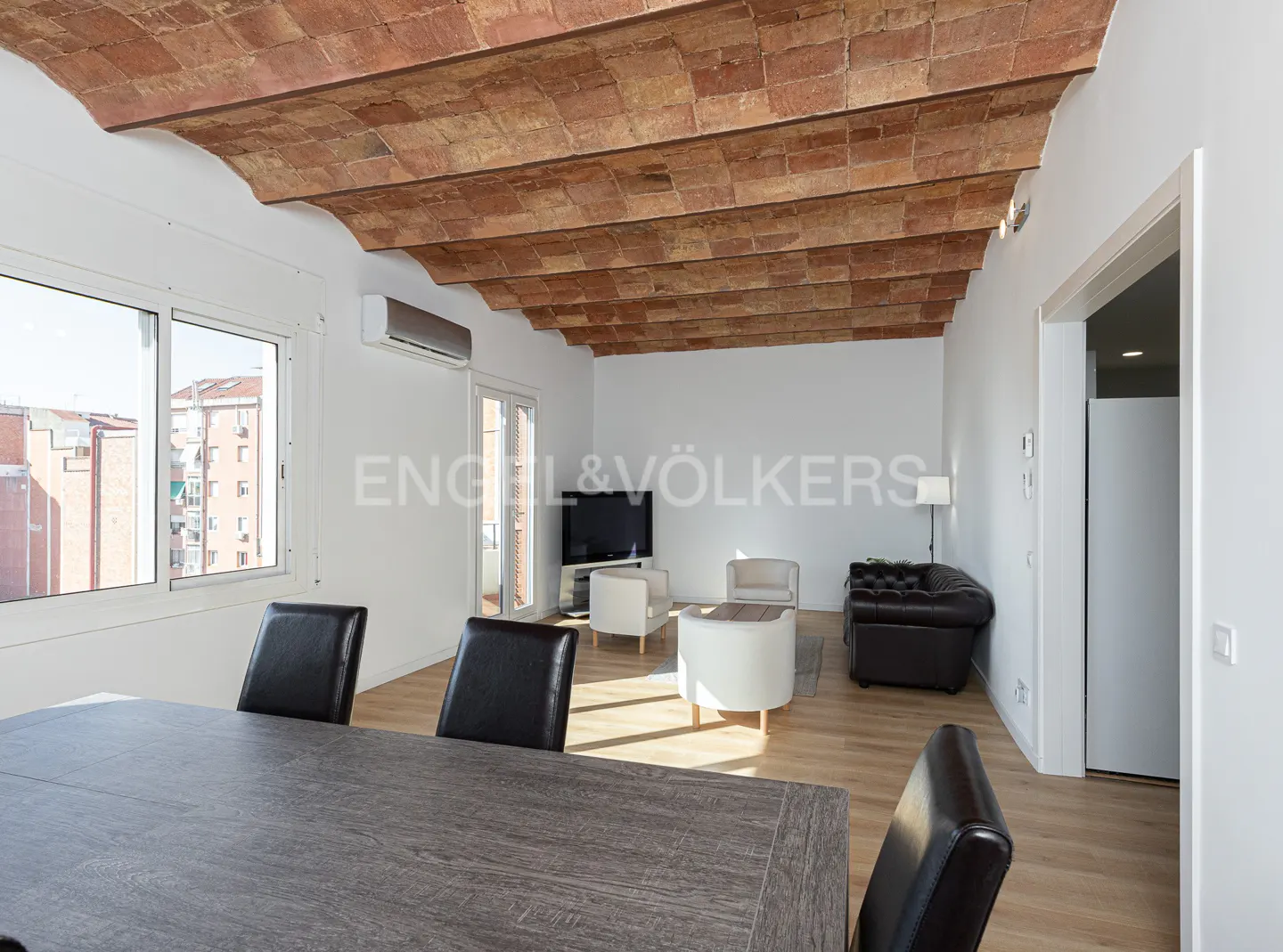 Bright living room with a brick vaulted ceiling, wood floors, and white walls. A gray table with black chairs is in the foreground. A brown leather sofa is in the background.