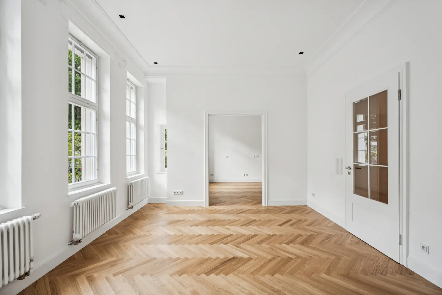 Bright, empty room with herringbone wood floors, white walls, windows, radiators, and a glass-paneled door.