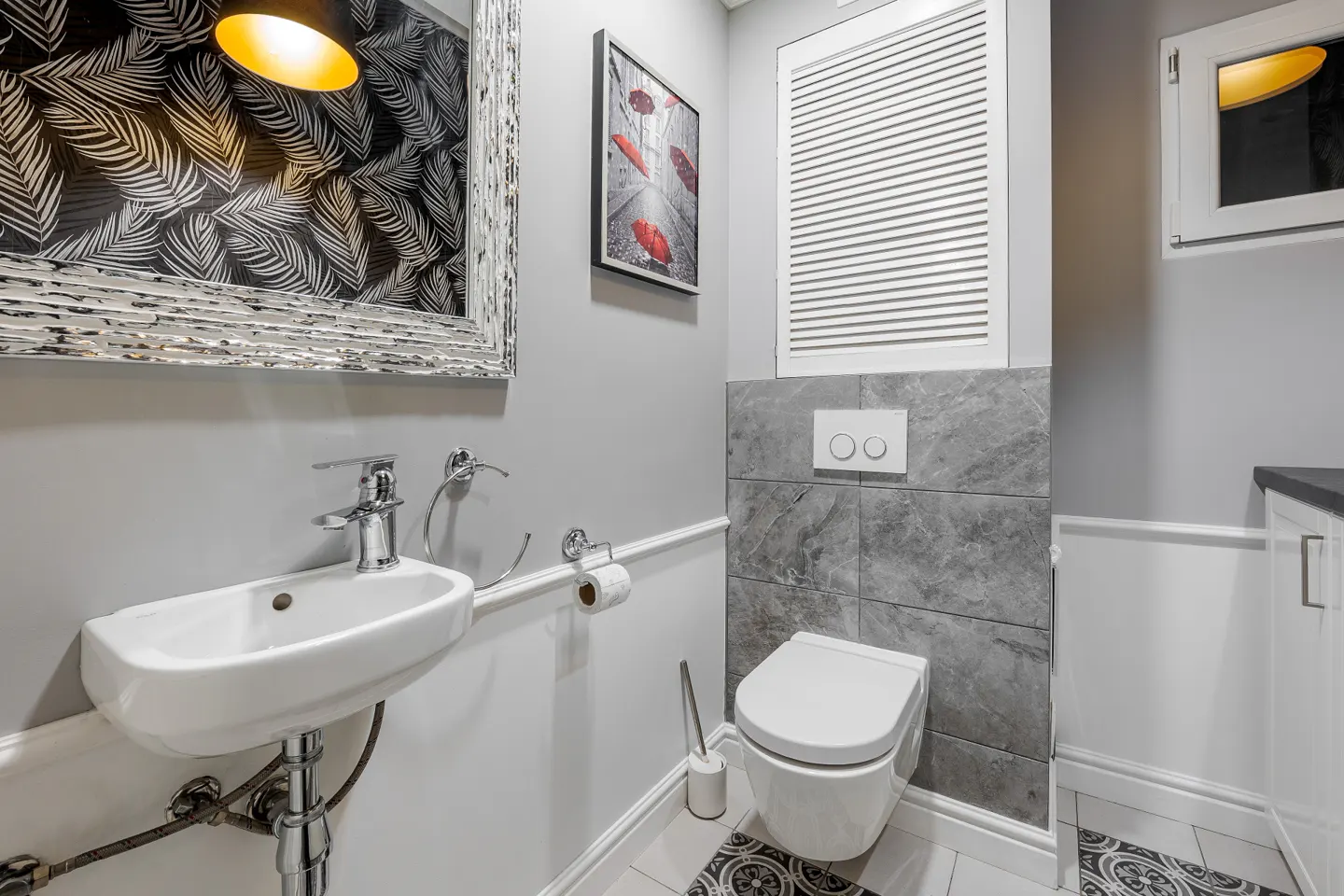 A small, gray powder room with a white sink, toilet, and gray marble tile. A mirror with a black and white leaf pattern hangs above the sink.