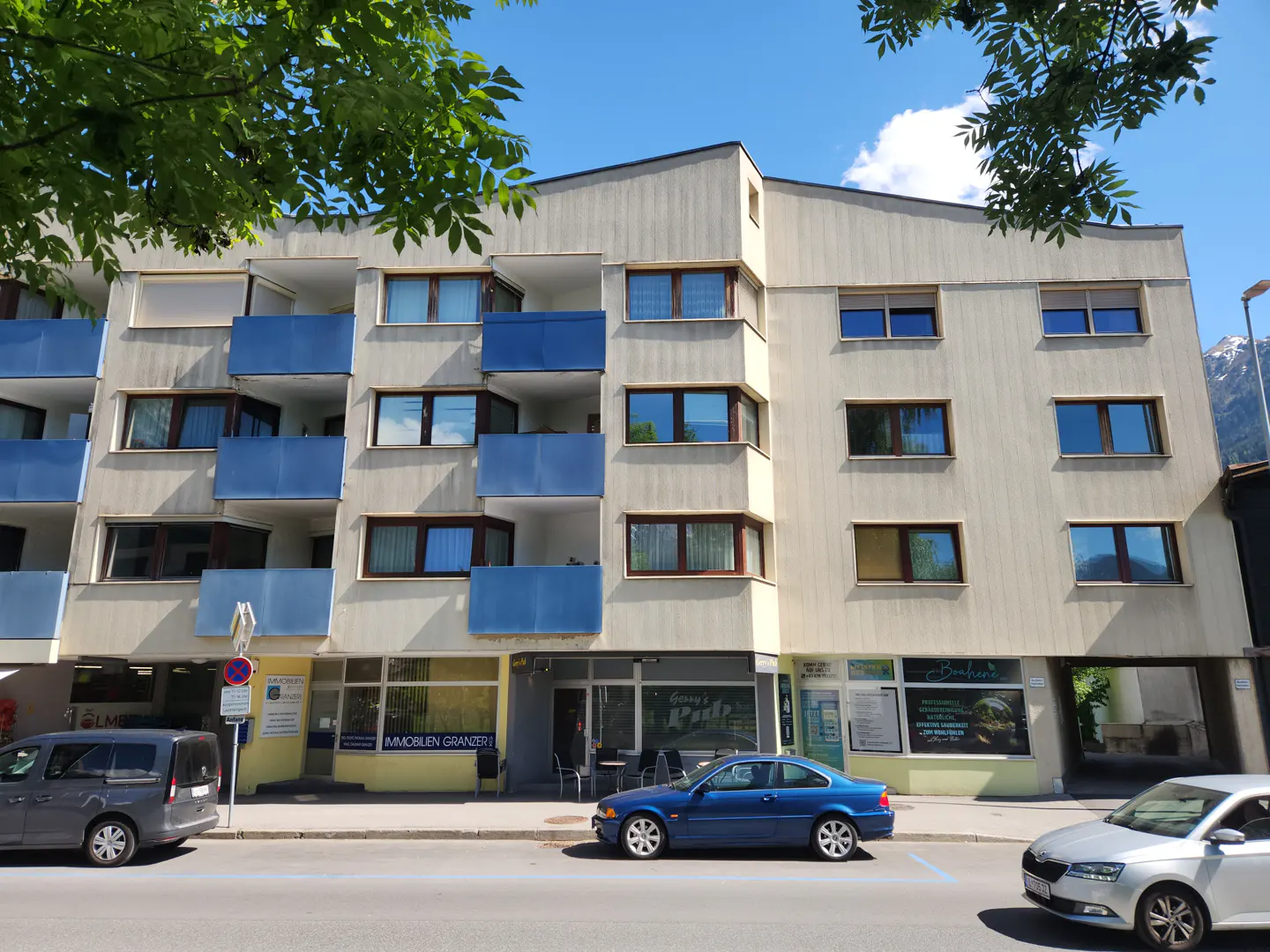 A four-story apartment building with blue balconies and street-level shops on a sunny day.