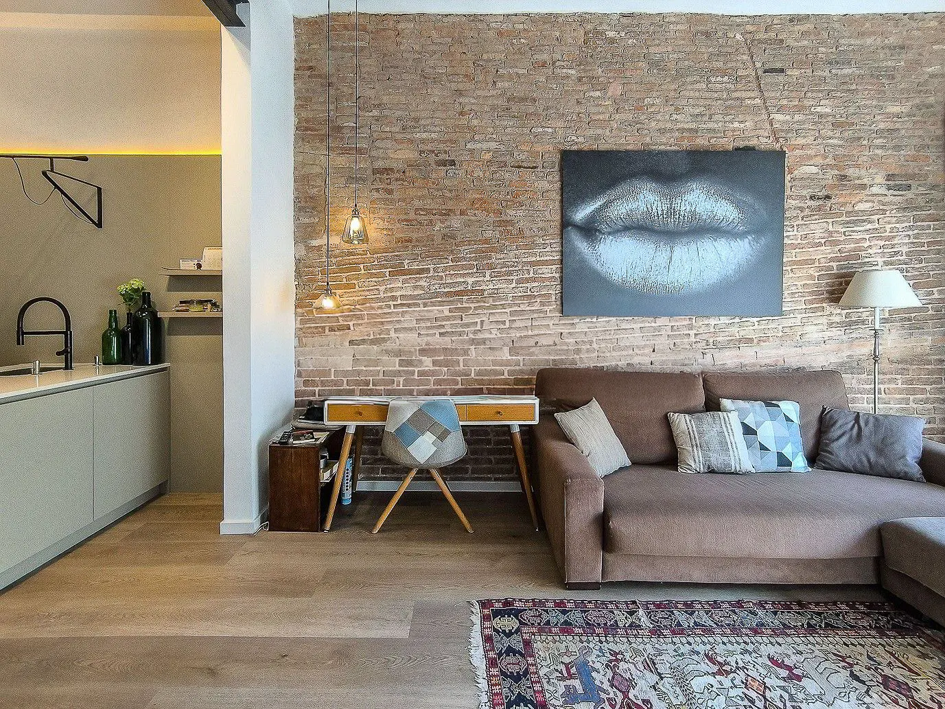 Living room with exposed brick wall, brown sofa, desk, and kitchen area. A large lip art hangs above the sofa.