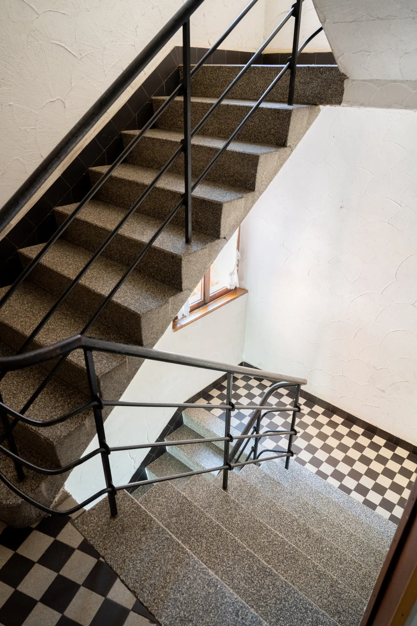 Staircase with grey stone steps, black metal railings, and a black-and-white checkered floor in a white-walled stairwell.