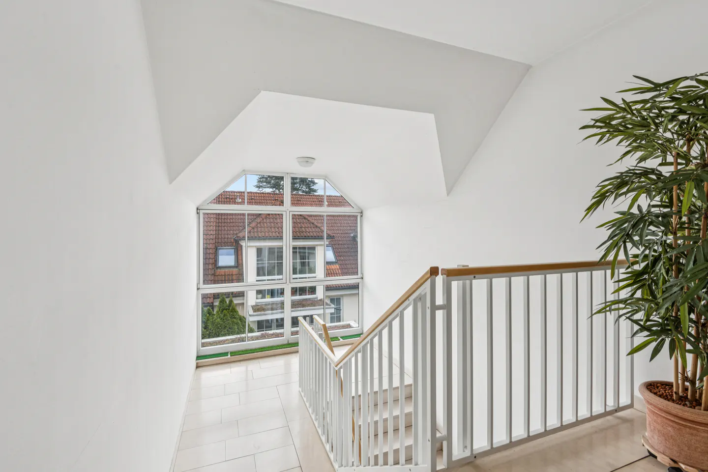 Bright hallway with white walls, tile floor, and stairs with white railing. Large window shows a house with a red tile roof. A potted plant is on the right.
