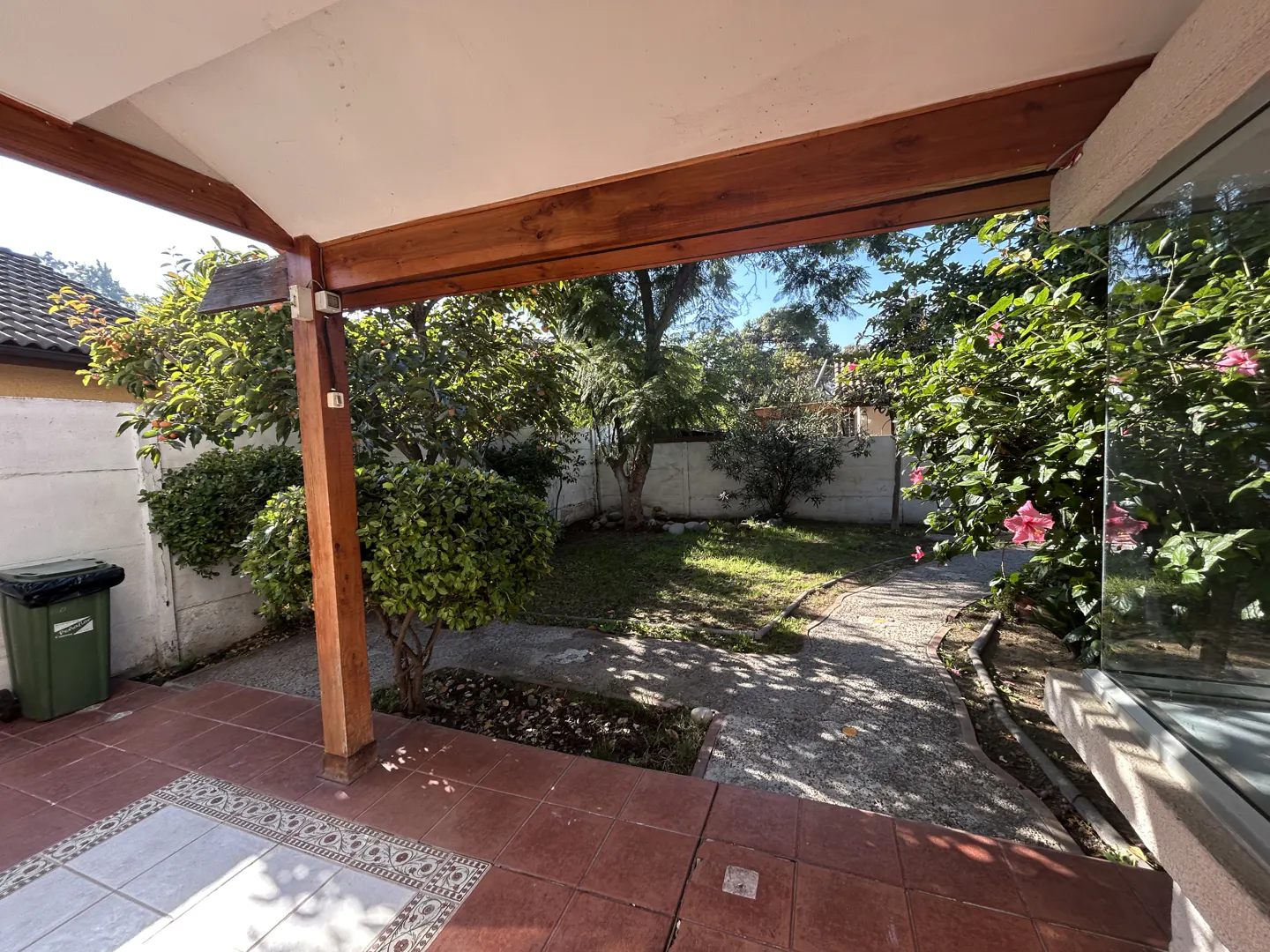Covered patio with red tile floor leads to a green lawn and garden with trees and a winding stone path.