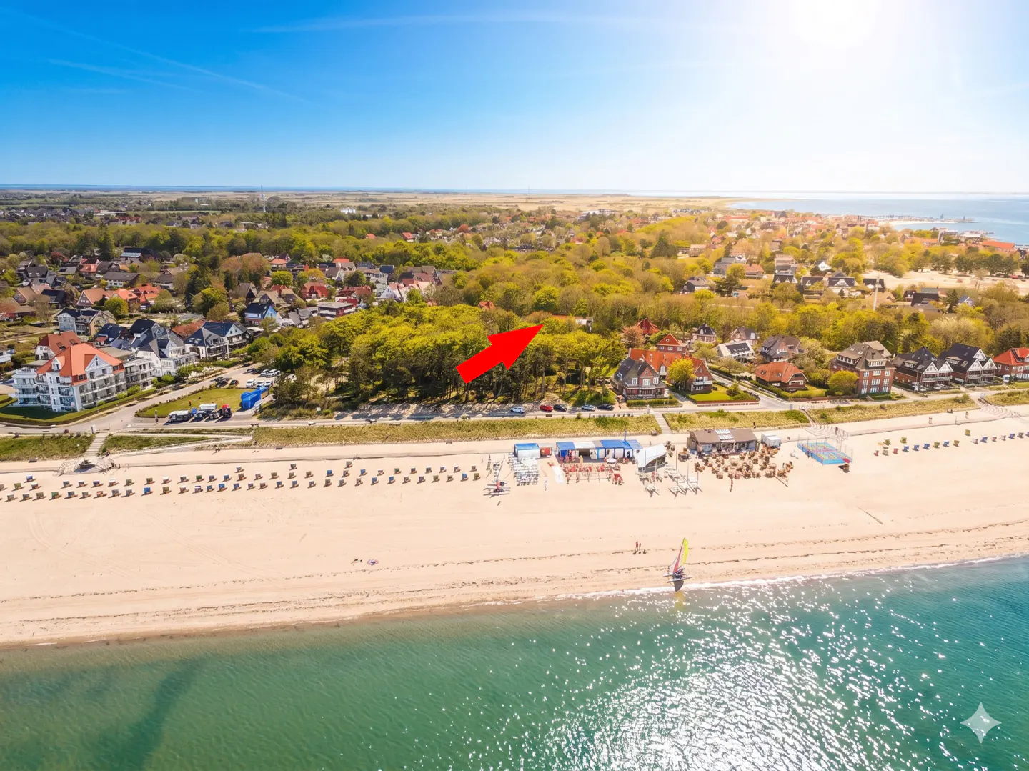 Aerial view of a sandy beach with blue water, beach chairs, and an arrow pointing to a house in the trees.