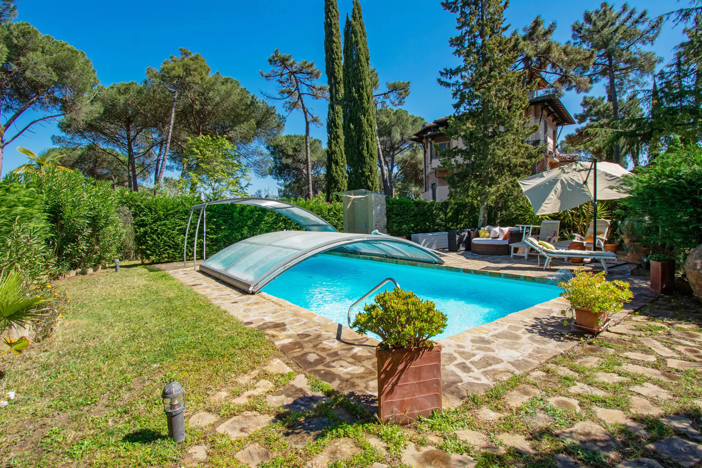 Outdoor pool with a retractable cover, surrounded by stone patio, green grass, trees, and lounge furniture.