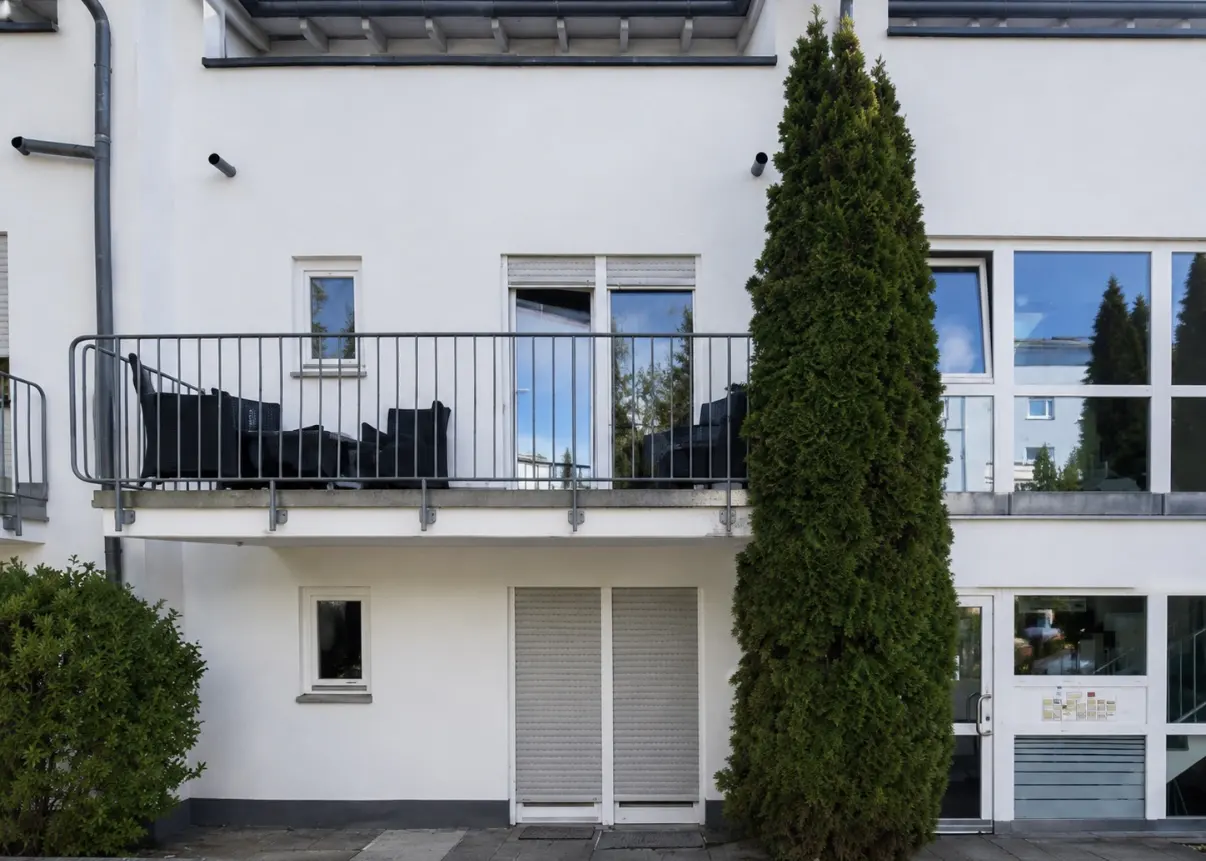 Exterior of a white building with a balcony, black patio furniture, and a tall green tree.